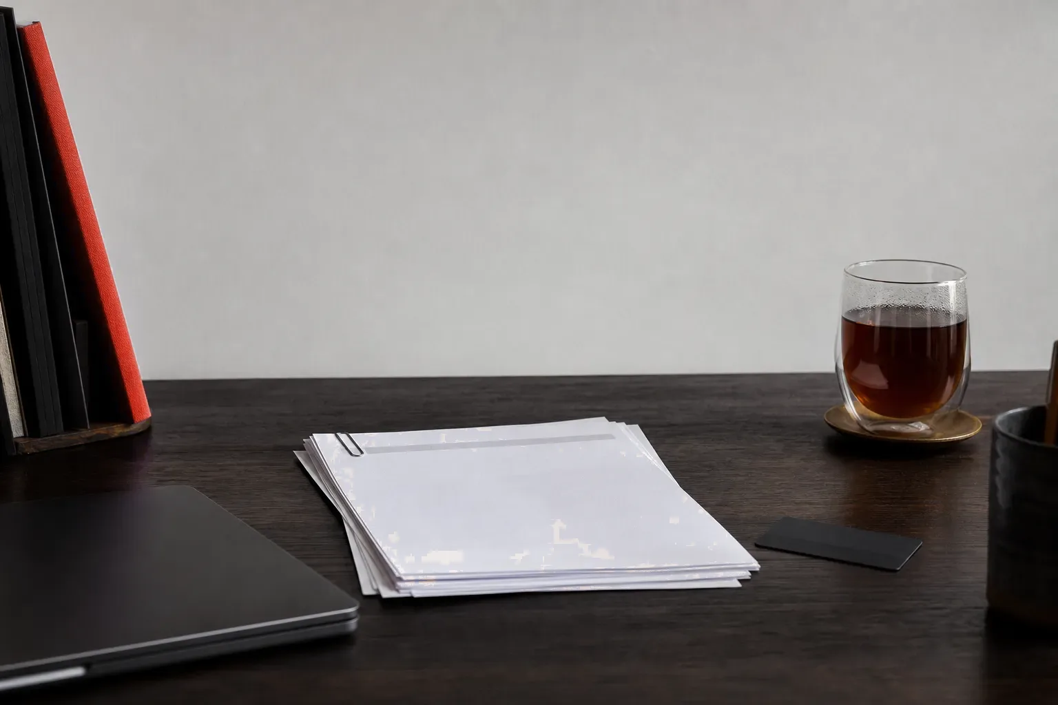A closed laptop, a clip of printed invoices and a red-spined book on a home-office desk
