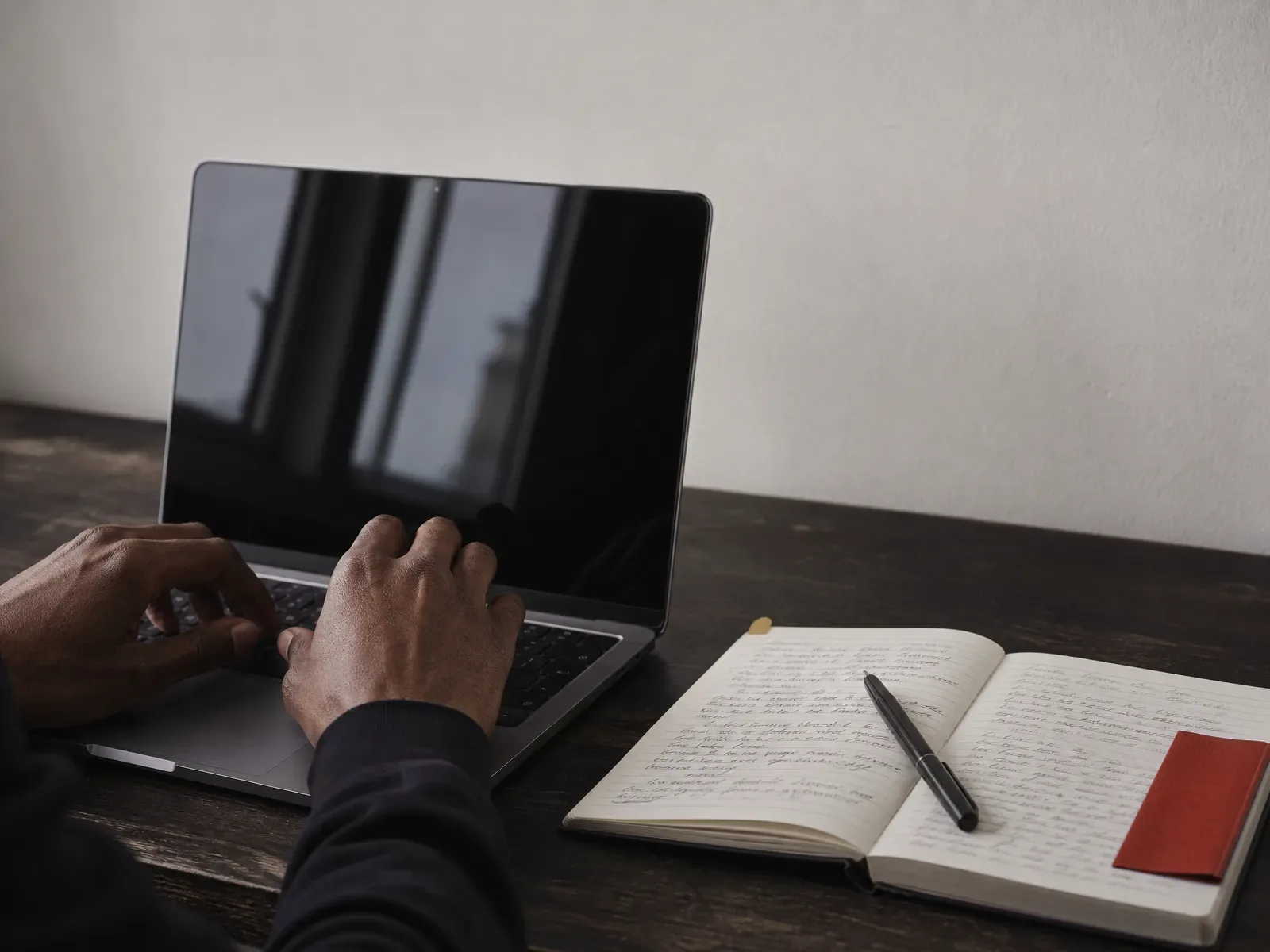 Hands resting on a laptop keyboard beside an open notebook with a red bookmark