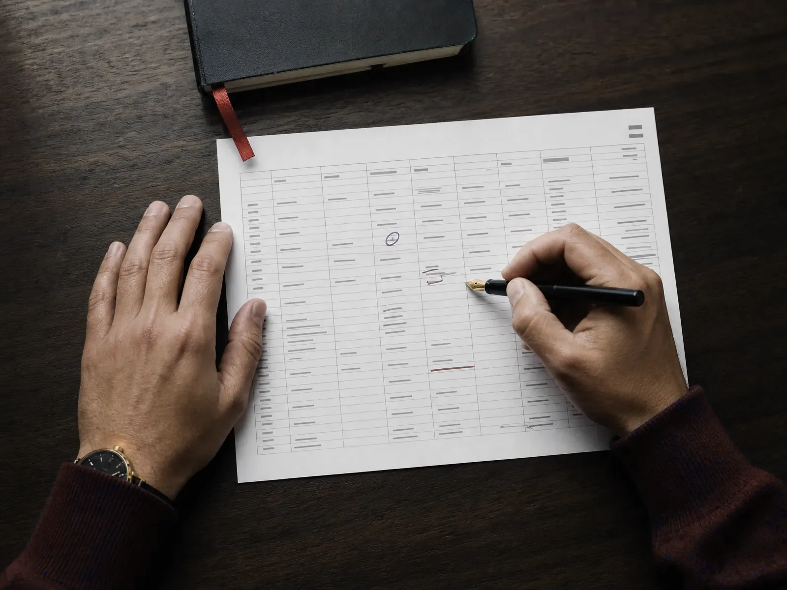 Hands annotating a printed spreadsheet with a fountain pen on a dark wood table