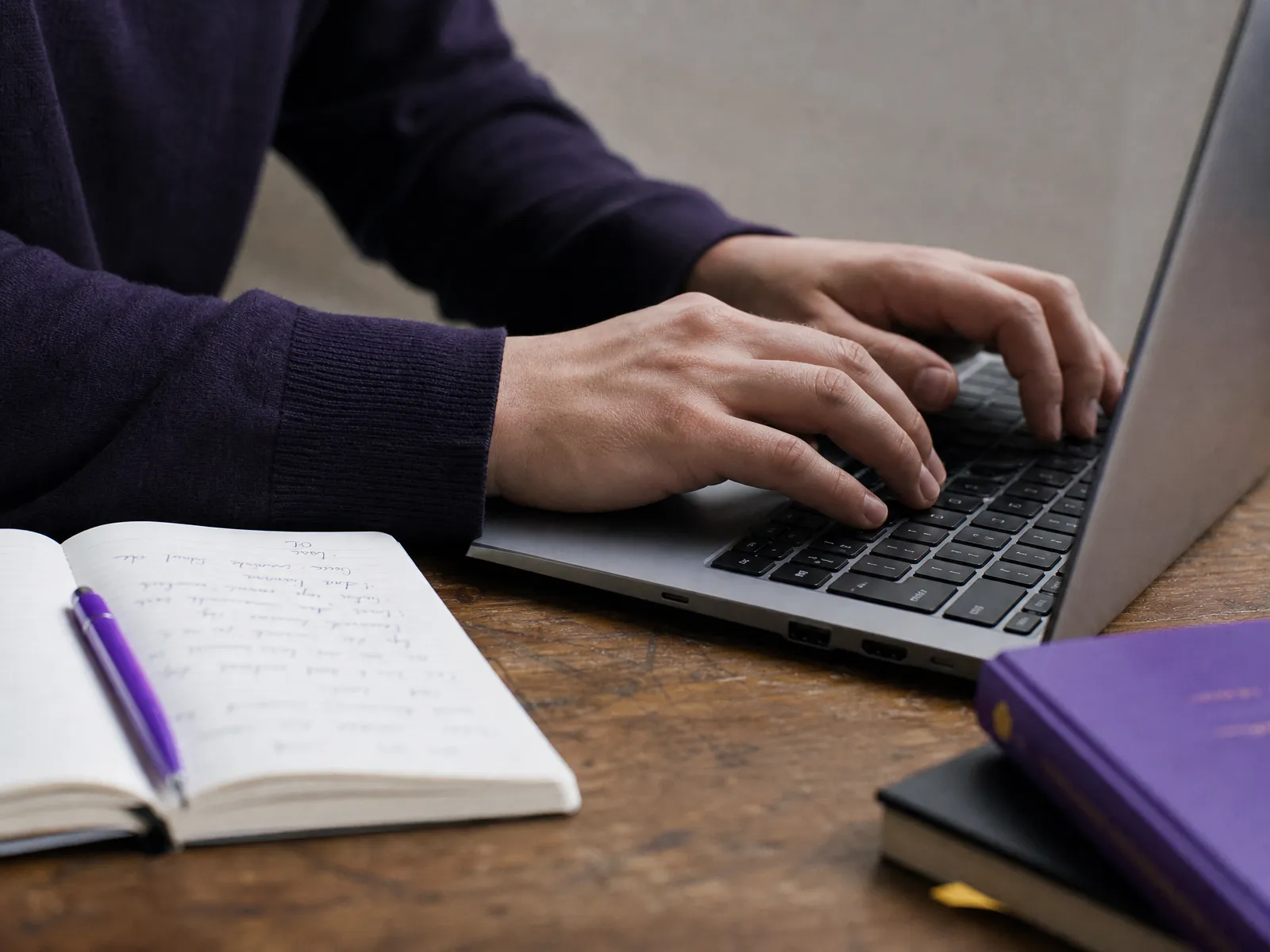 Hands resting on a laptop keyboard beside an open notebook on a wooden desk in soft daylight, working through blog SEO.