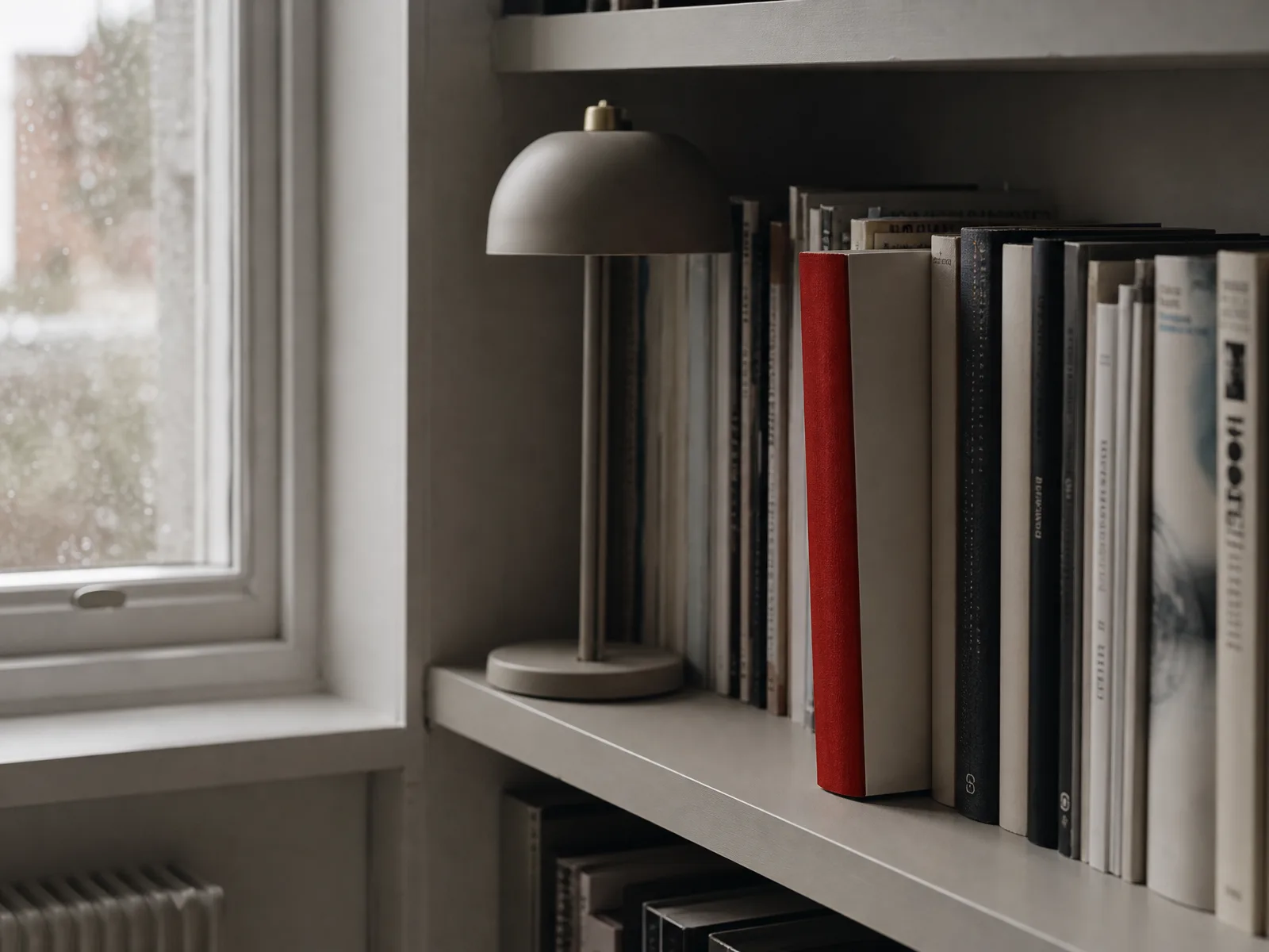 A red-spined book on a shelf beside a window and a small lamp