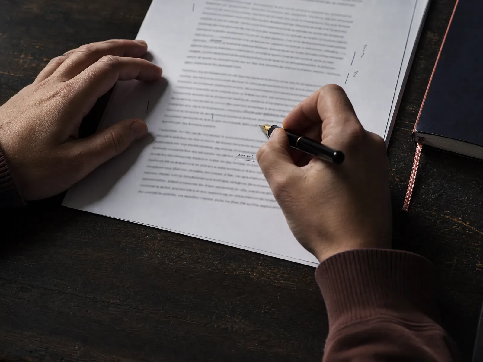 Hands annotating a printed page with a fountain pen beside a notebook with a red ribbon