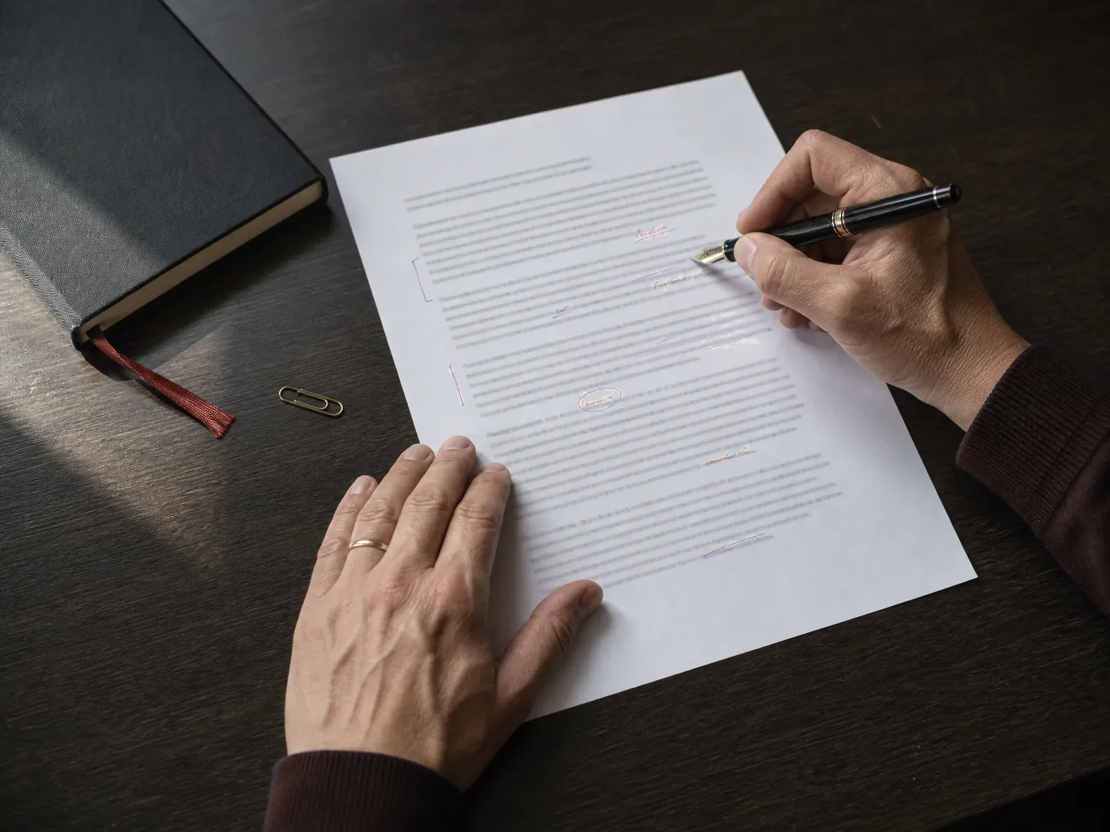 Hands annotating a printed document with a fountain pen beside a notebook with a red bookmark