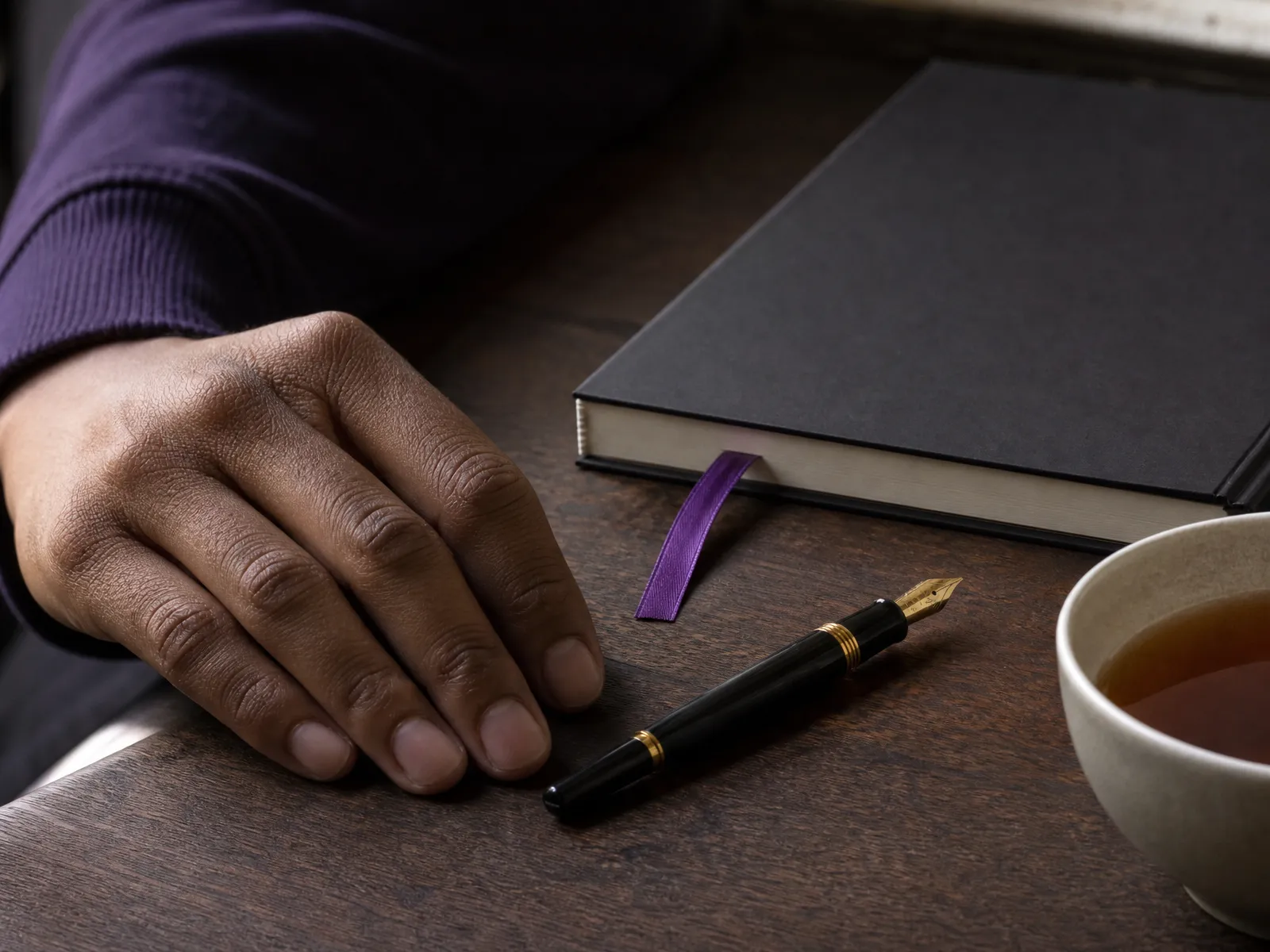 Close crop of a hand beside a fountain pen on a dark wood desk, a violet ribbon bookmark in a closed notebook.