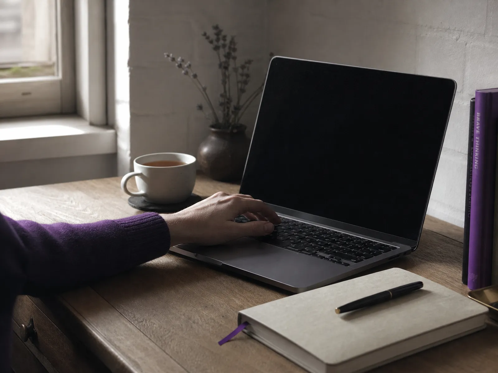 A hand on a laptop keyboard at a tidy desk, a notebook and pen alongside.