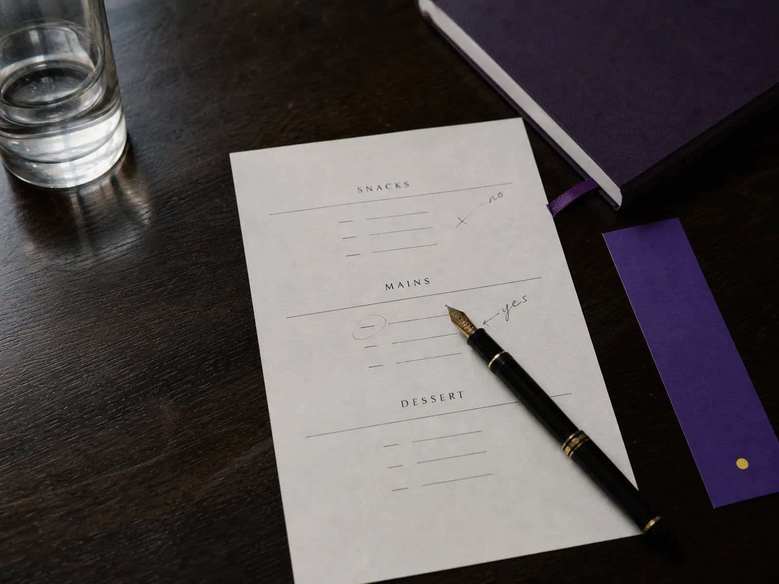 A printed restaurant menu on a wooden table with a fountain pen and a violet bookmark.