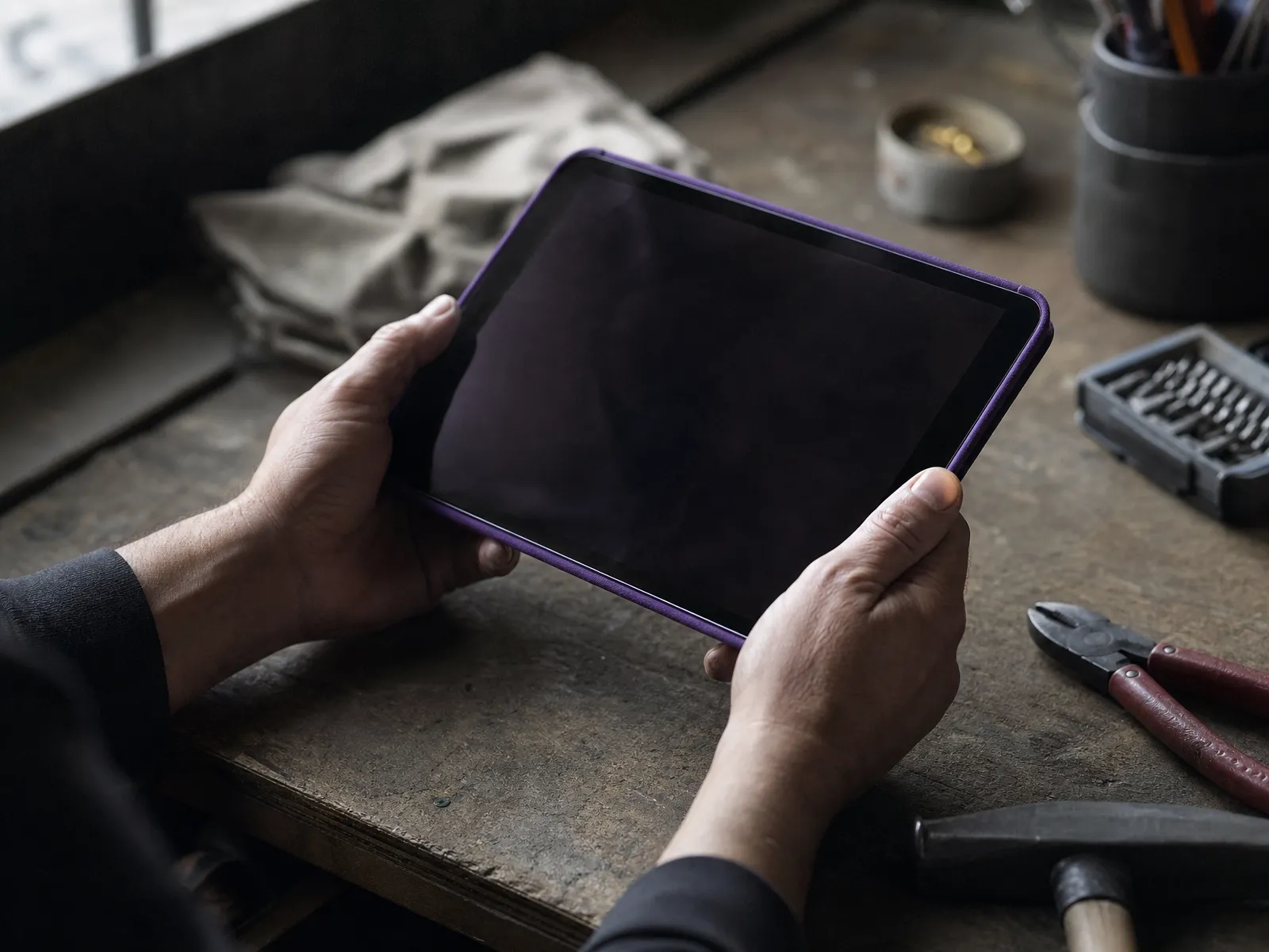 Hands holding a tablet with a dark screen at a cluttered tradesman's workbench.