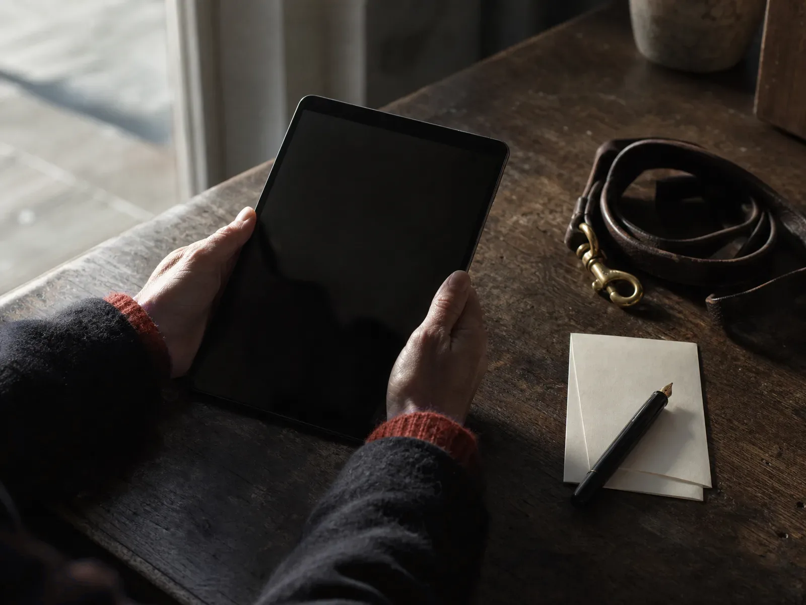 Hands holding a tablet with a dark screen on a wooden counter beside a coiled leather
