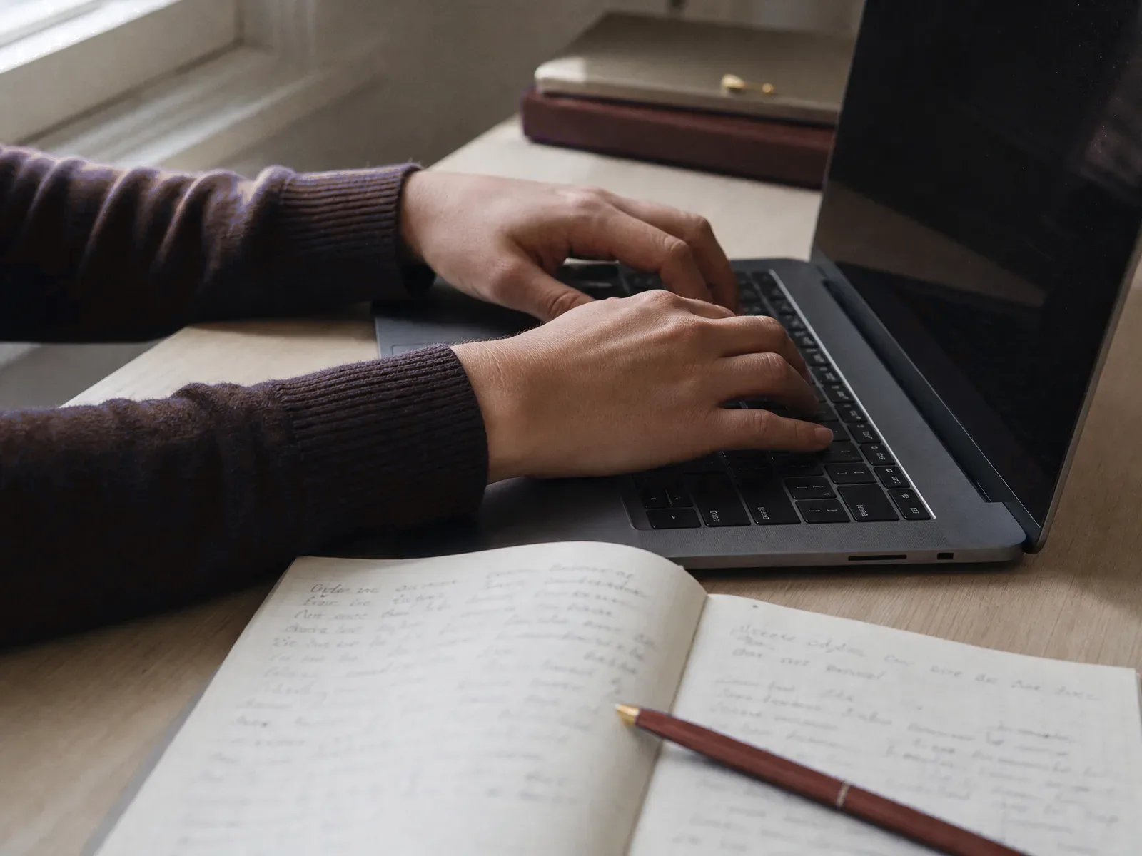 Hands typing on a laptop beside an open notebook and red pen