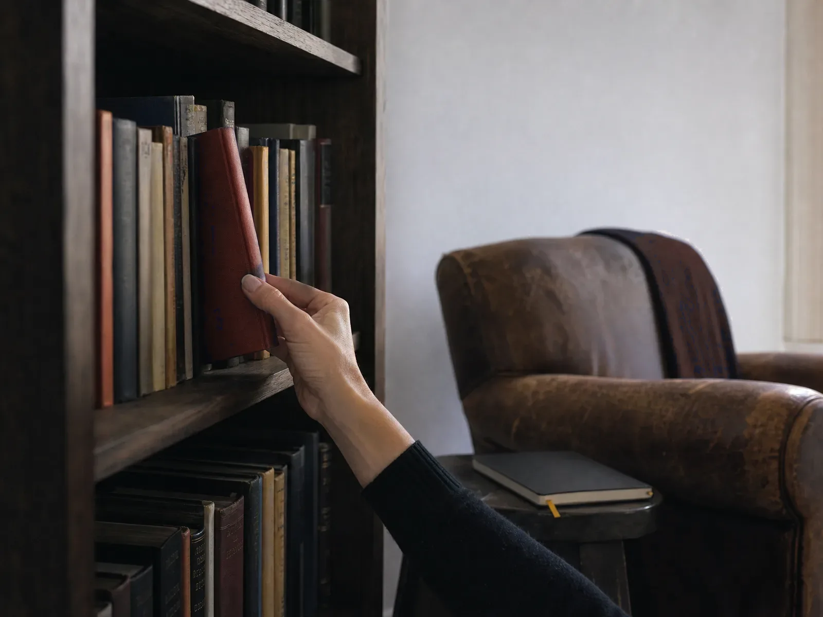 Hand lifting a red-spined book from a dark bookshelf beside a leather chair