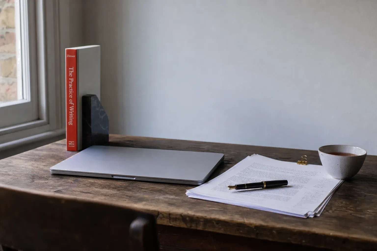 A wooden desk with a closed laptop, printed pages and a fountain pen