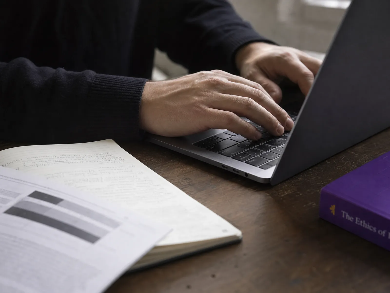 Hands resting on a laptop keyboard beside a printed page and an open notebook, in soft daylight on a wooden desk.