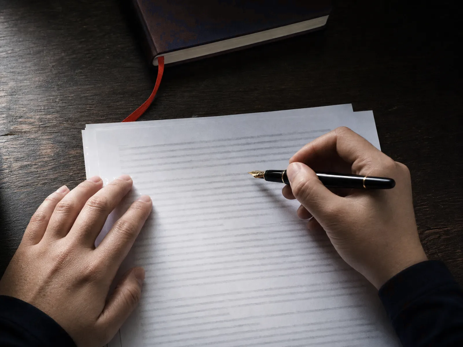 Two hands annotating a stack of printed pages with a fountain pen on dark wood