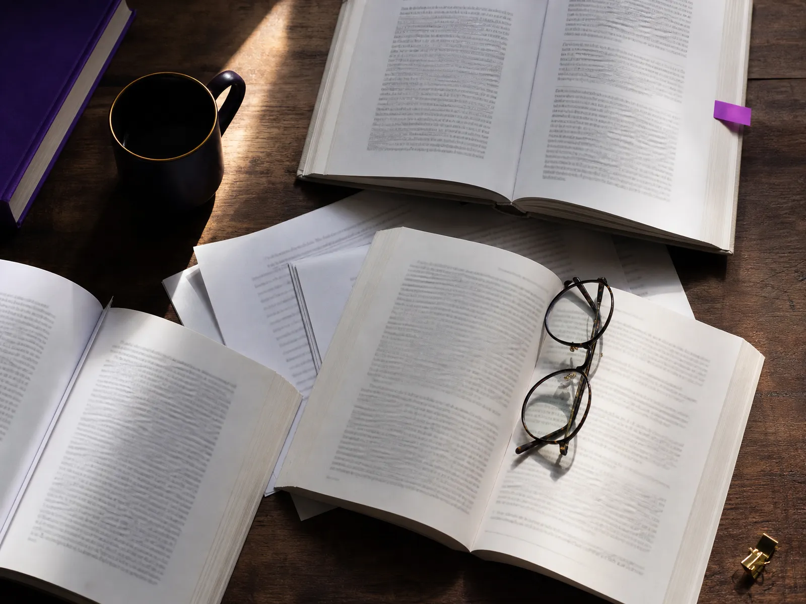 Open books and printed pages fanned out with reading glasses on a desk