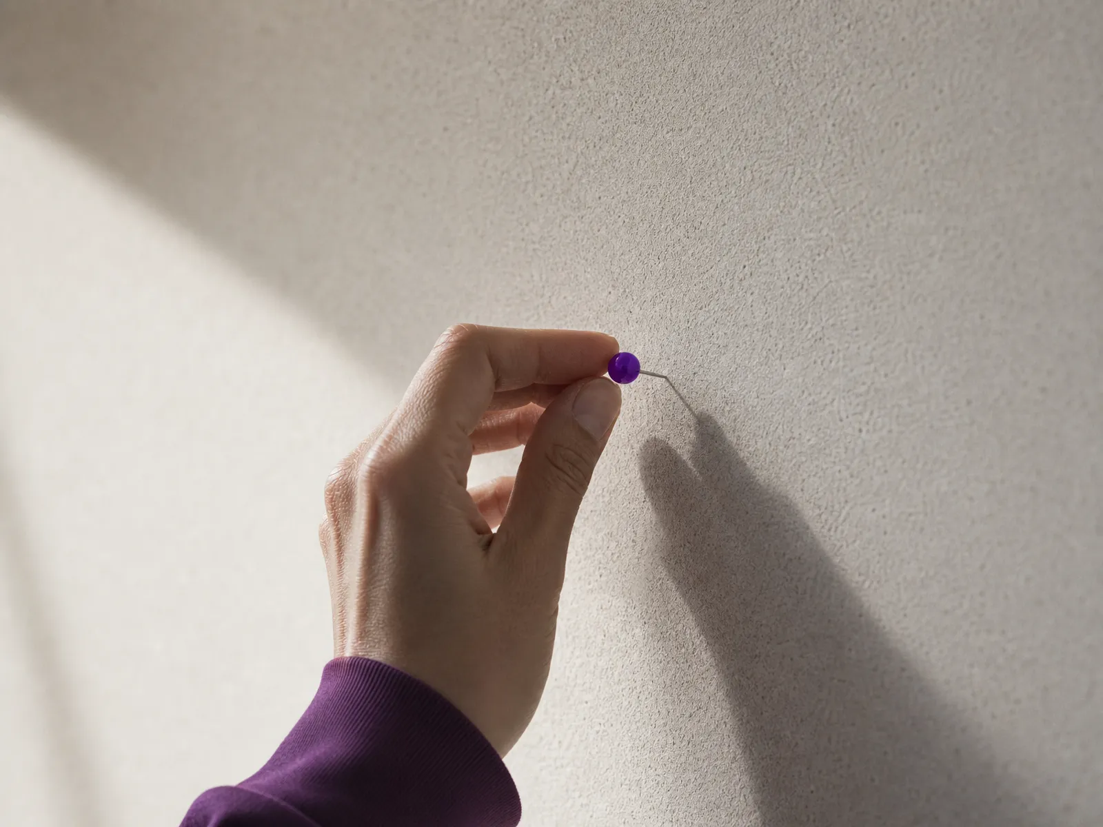 A hand pressing a single violet pin into a plain corkboard