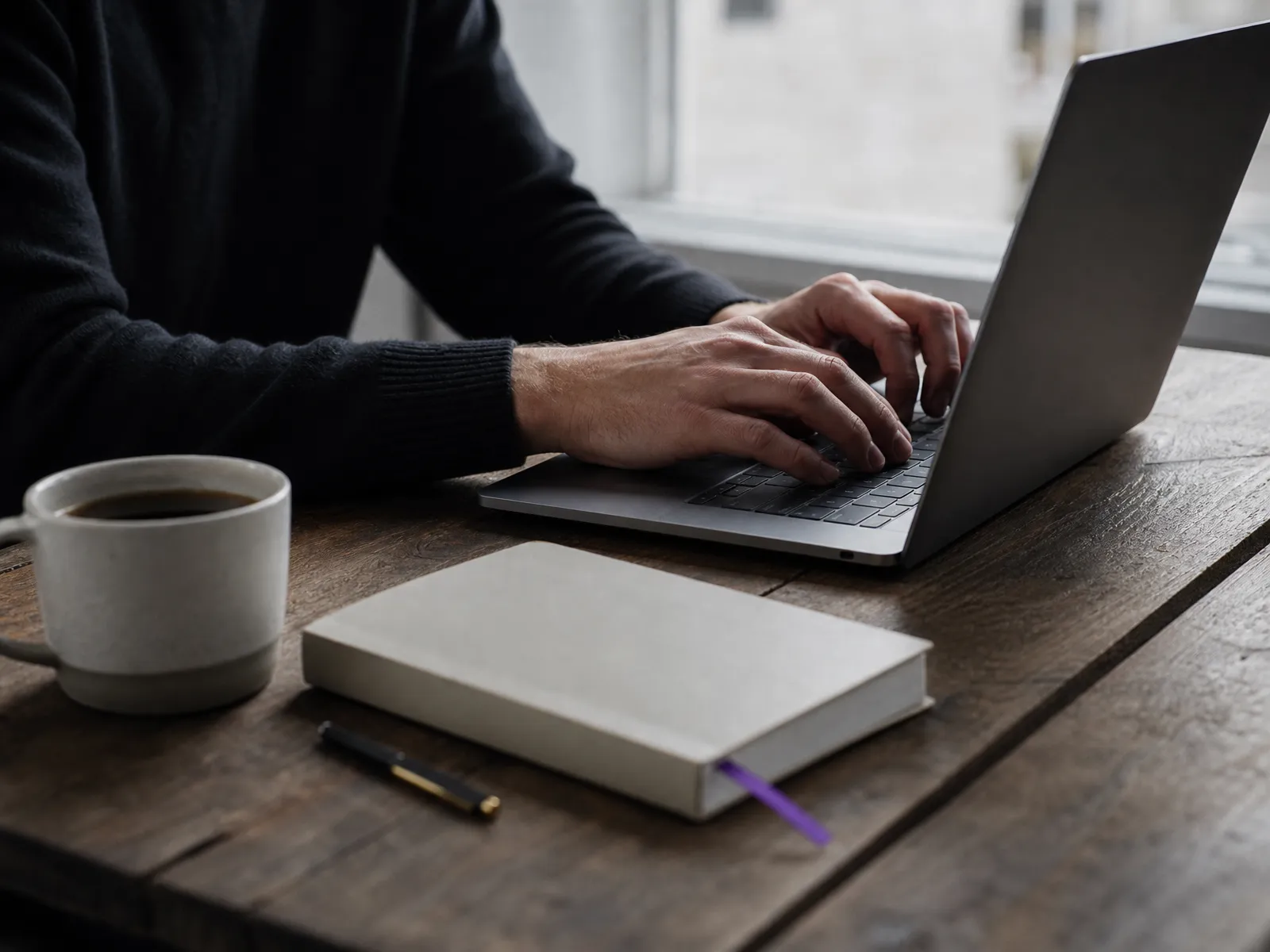 Hands working on a laptop with a notebook and coffee, screen turned away